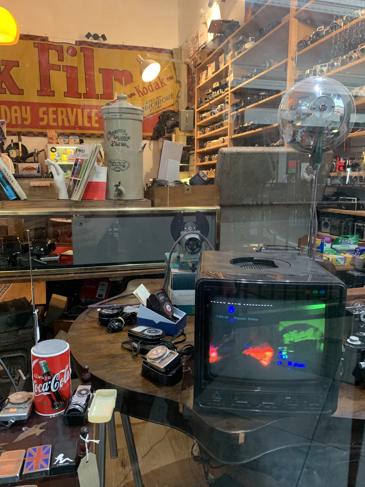 A vintage camera shop window displaying Kodak film signs, a disco ball and a CRT television showing a retro game