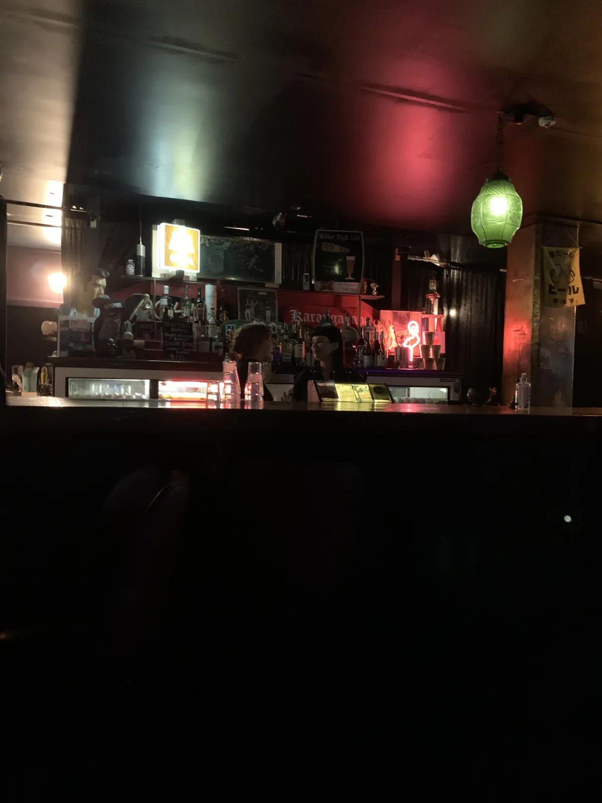 The dimly lit interior of a K Road bar, green lantern above the counter, a Karangahape Road neon sign glowing red behind the bar