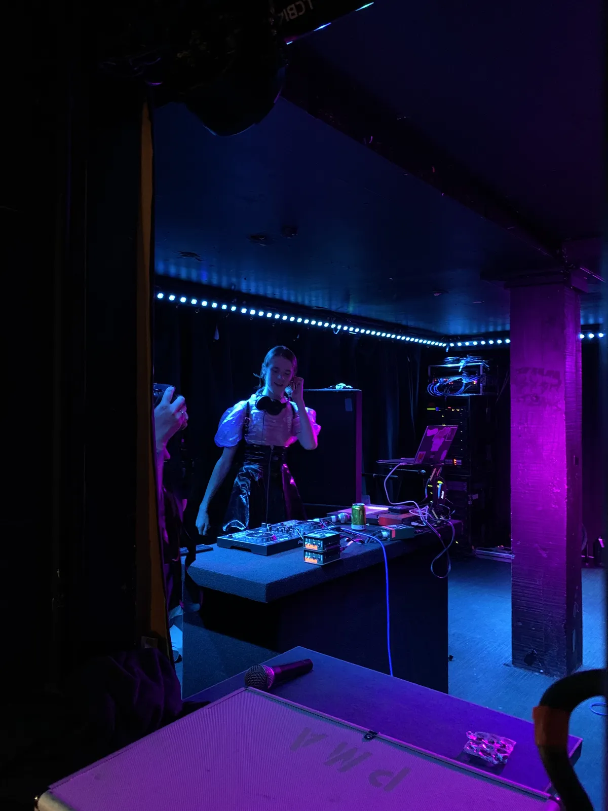A DJ performing under blue-purple stage lighting in a K Road venue, headphones around her neck, mixer in front