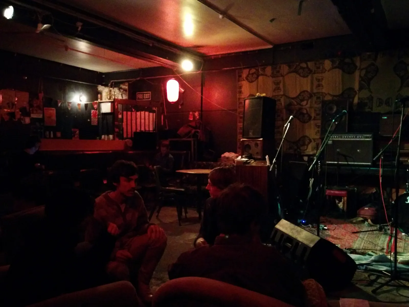 A packed audience in a red-lit basement venue, Japanese paper lantern hanging near the fire exit sign, walls covered in posters