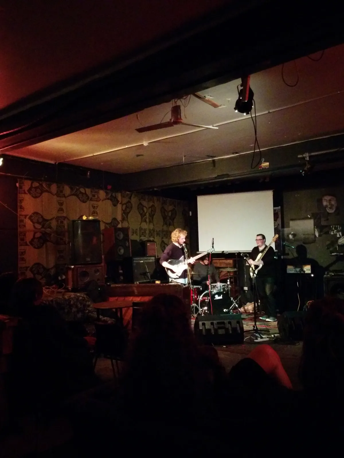 A band playing a live set in a raw K Road venue in 2014, patterned wallpaper, a projection screen behind the drummer